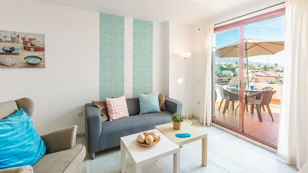 Un salon moderne avec un canapé gris, une petite table blanche sur laquelle se trouve un saladier rempli de fruits, et un balcon agrémenté de mobilier d’extérieur.