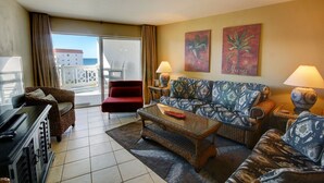 Une chambre d’hôtel avec un balcon, un fauteuil rouge, un canapé à motifs, une table basse et deux lampes.