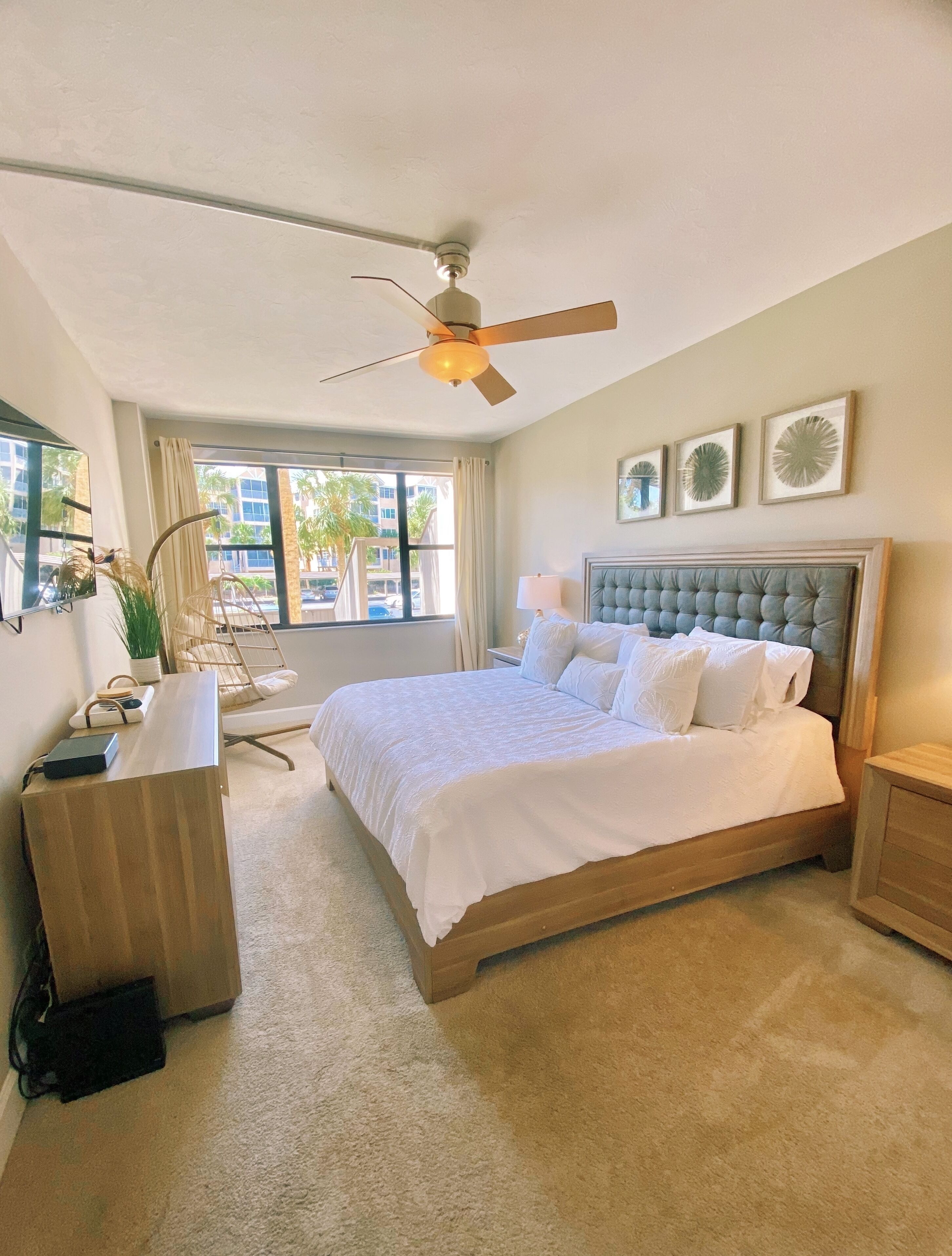 Un dormitorio con cama, escritorio, silla, ventilador de techo y ventana con vistas a palmeras y edificios.