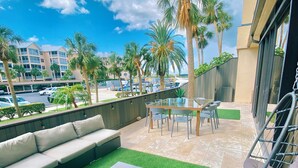Um terraço no telhado com um sofá, uma mesa e cadeiras. Há palmeiras e uma vista de um prédio de hotel ao fundo.