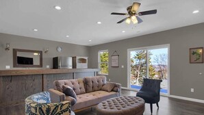 A spacious living room with a sofa, ottoman, and armchair, a ceiling fan, and a kitchen area with a TV. - The Bittersweet Inn: Brand new 6 bed 5 bath house (Hermann)
