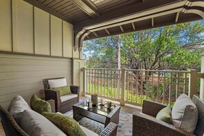 A covered outdoor seating area with wicker furniture, a glass table, and a view of trees.