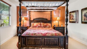 A four-poster bed with a floral bedspread, flanked by two nightstands with lamps, in a room with a window and artwork on the wall.