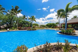 A clear blue pool surrounded by palm trees and tropical plants, with a gazebo and lounge chairs in the background.