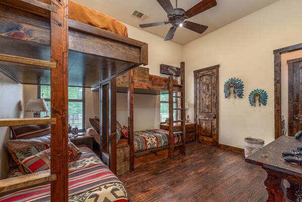 Habitación rústica con cama de altillo, litera inferior y una cama tipo diván. Hay una mesa de madera con una cesta y un ventilador de techo.