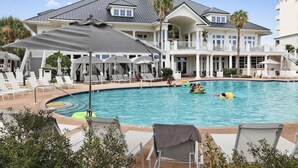 A large swimming pool with lounge chairs and umbrellas, surrounded by a large house with a balcony.