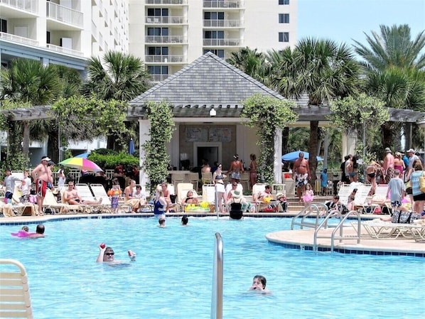 A pool area with lounge chairs and umbrellas, surrounded by palm trees and a building with a tiled roof.