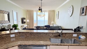 A modern kitchen with granite countertops, a dishwasher, and a sink. There is a dining area with a sofa and a view of the ocean.