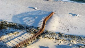 A beach with a wooden boardwalk leading to the water, a sign with 'MELBOURNE' written on the sand, and a clear view of the ocean waves.