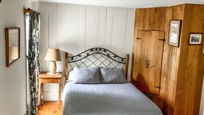 Un dormitorio con piso de madera, una cama con cabecera, una mesita de noche, una lámpara y un armario de madera.