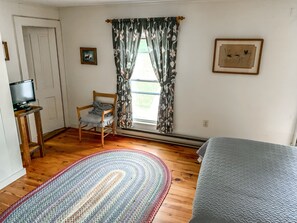 A room with a wooden floor, a bed, a chair, a television on a stand, and a window with curtains.