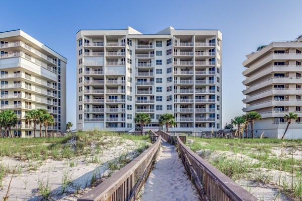 A beachfront hotel with multiple floors, balconies, and a sandy walkway leading to the beach.
