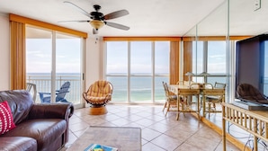 A spacious living area with a large window overlooking the ocean, a ceiling fan, a dining table with chairs, and a leather sofa. - Toes In the Sand!!! (Pensacola)