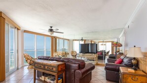 A spacious living room with a large brown leather sofa, a wooden coffee table, and a television. There is a balcony with large windows and a view of the ocean. - Toes In the Sand!!! (Pensacola)