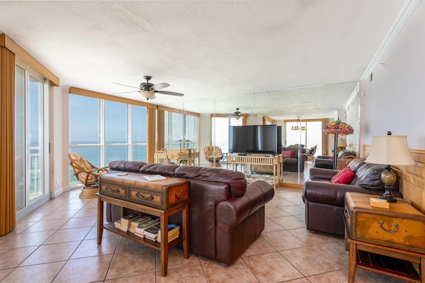 A spacious living room with a large brown leather sofa, a wooden coffee table, and a television. There is a balcony with large windows and a view of the ocean. - Toes In the Sand!!! (Pensacola)