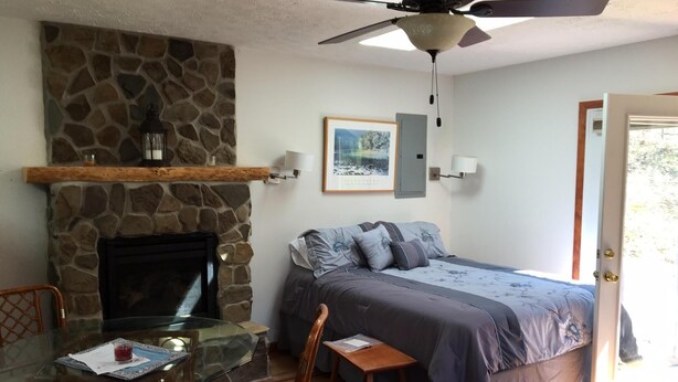 Une chambre avec une cheminée en pierre, un lit recouvert d’une couvre-lit bleue et grise, une table en verre avec des chaises et un ventilateur de plafond.