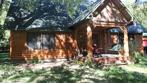 A log cabin with a porch, a swing, and a window overlooking a wooded area. - Cozy, Family Friendly Log Cabin Close To Everything! (Williams Bay)