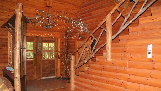 A log cabin interior with a wooden staircase, a chandelier, and a door leading outside. - Cozy, Family Friendly Log Cabin Close To Everything! (Williams Bay)