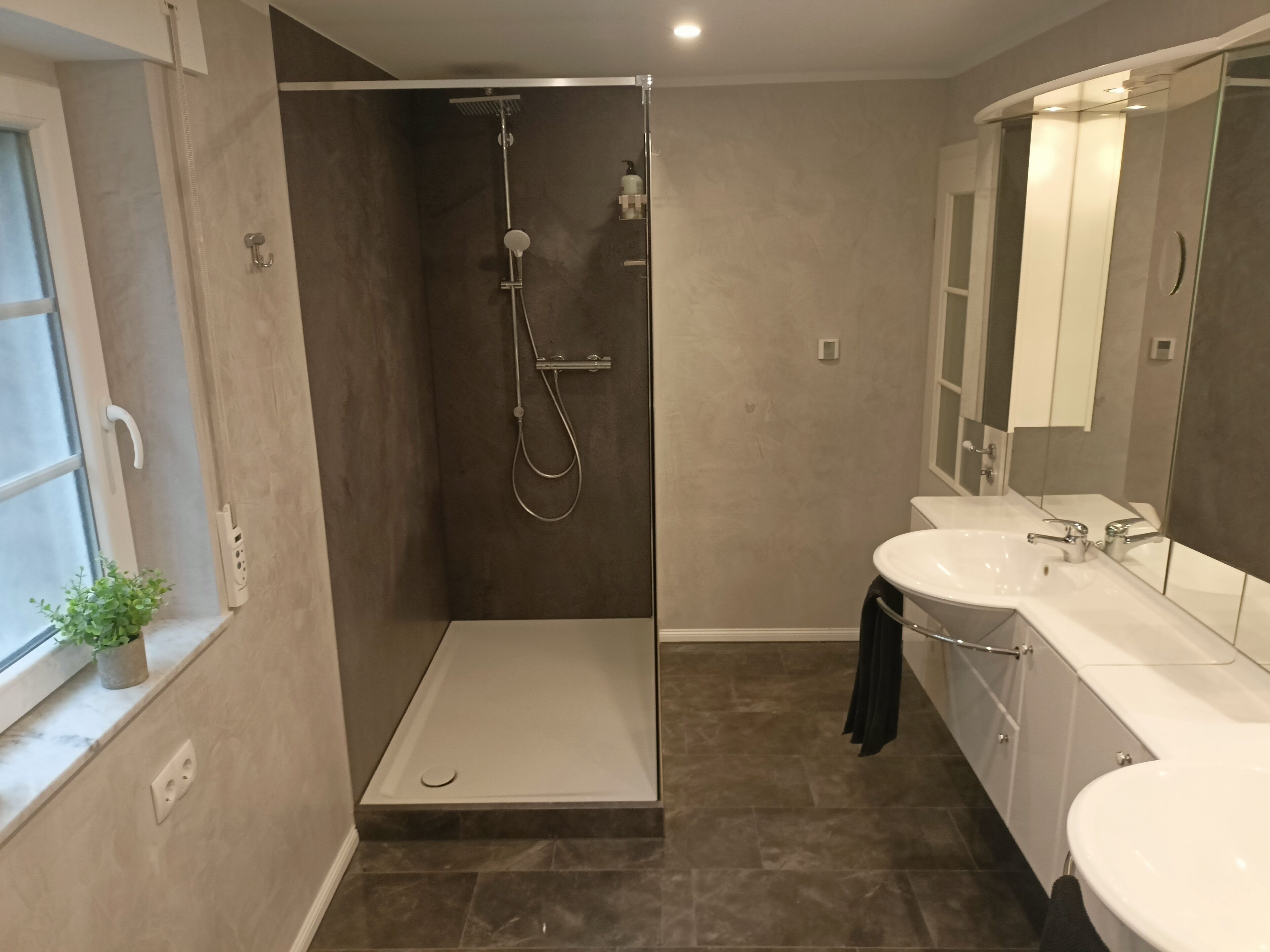 A modern bathroom with a walk-in shower, a vanity with a mirror, and a window with a plant.