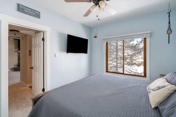 A bedroom with a bed, a ceiling fan, a TV, and a window with a view of trees. - Après-Ski Hideaway | Ski Locker & Hot Tubs (Steamboat Springs)