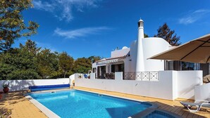 Une piscine avec une terrasse ensoleillée, un bâtiment blanc doté d’une cheminée et un ciel d’un bleu limpide.