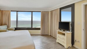 Ett hotellrum med en stor säng, en tv på ett stativ, utsikt över havet och ett fönster med gardiner.