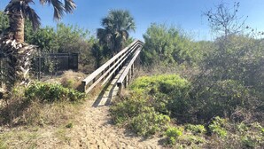 A sandy path leading to a wooden bridge over a fence, surrounded by greenery and palm trees. - GOIN COASTAL Beach House, 3/2, Sleeps 10, 3 min Walk To The Beach  (St. Augustine)