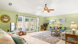 A spacious living room with a ceiling fan, a large window, a television, and a variety of seating options including a sofa and chairs. - GOIN COASTAL Beach House, 3/2, Sleeps 10, 3 min Walk To The Beach  (St. Augustine)