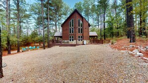 A log cabin with a large front porch and a gravel driveway, surrounded by tall pine trees. - Yellow Rose Cabin: Pet-Friendly Hochatown Getaway (sleeps 12) w/ Hot Tub, Arcade (Broken Bow)