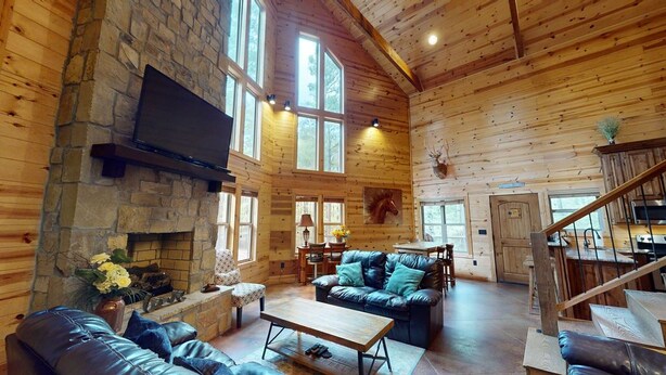A spacious log cabin interior with a stone fireplace, leather furniture, a television mounted on the wall, and a wooden staircase. - Yellow Rose Cabin: Pet-Friendly Hochatown Getaway (sleeps 12) w/ Hot Tub, Arcade (Broken Bow)