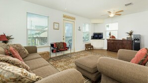 A spacious living room with a large sofa, ottoman, and a television. There is a kitchen area with a refrigerator and a dining table. - Phoenix Rising - Chill Beach Vibes, 4 Decks! (Surfside Beach)