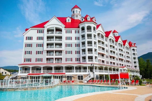 A multi-story hotel with a red roof, balconies, and a swimming pool area with sun loungers.