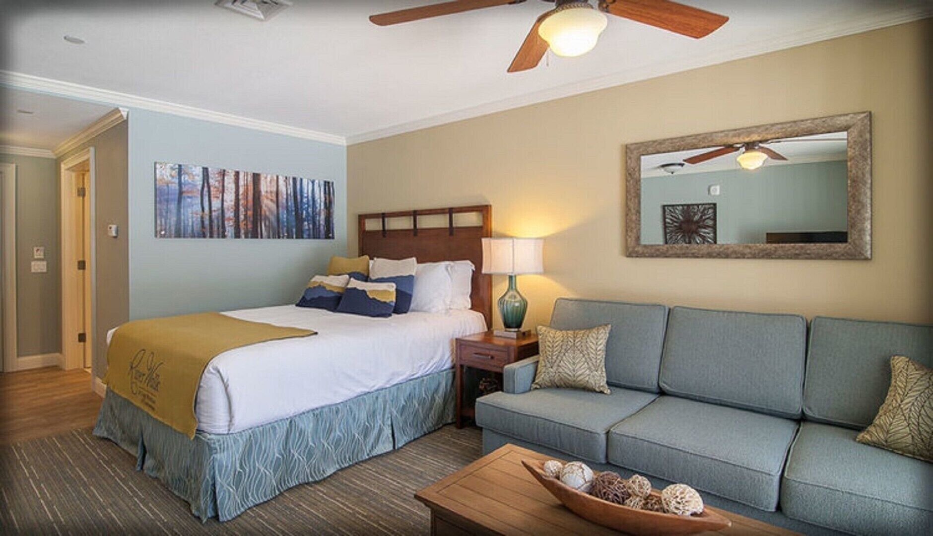 A hotel room with a bed, a sofa, a coffee table with decorative items, a lamp, a ceiling fan, and a large mirror on the wall.