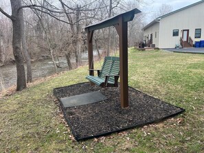 A wooden swing set with benches, a covered area, and a building in the background. - Creek Side Home-Fly Fishing, Nature Walks (Boiling Springs)