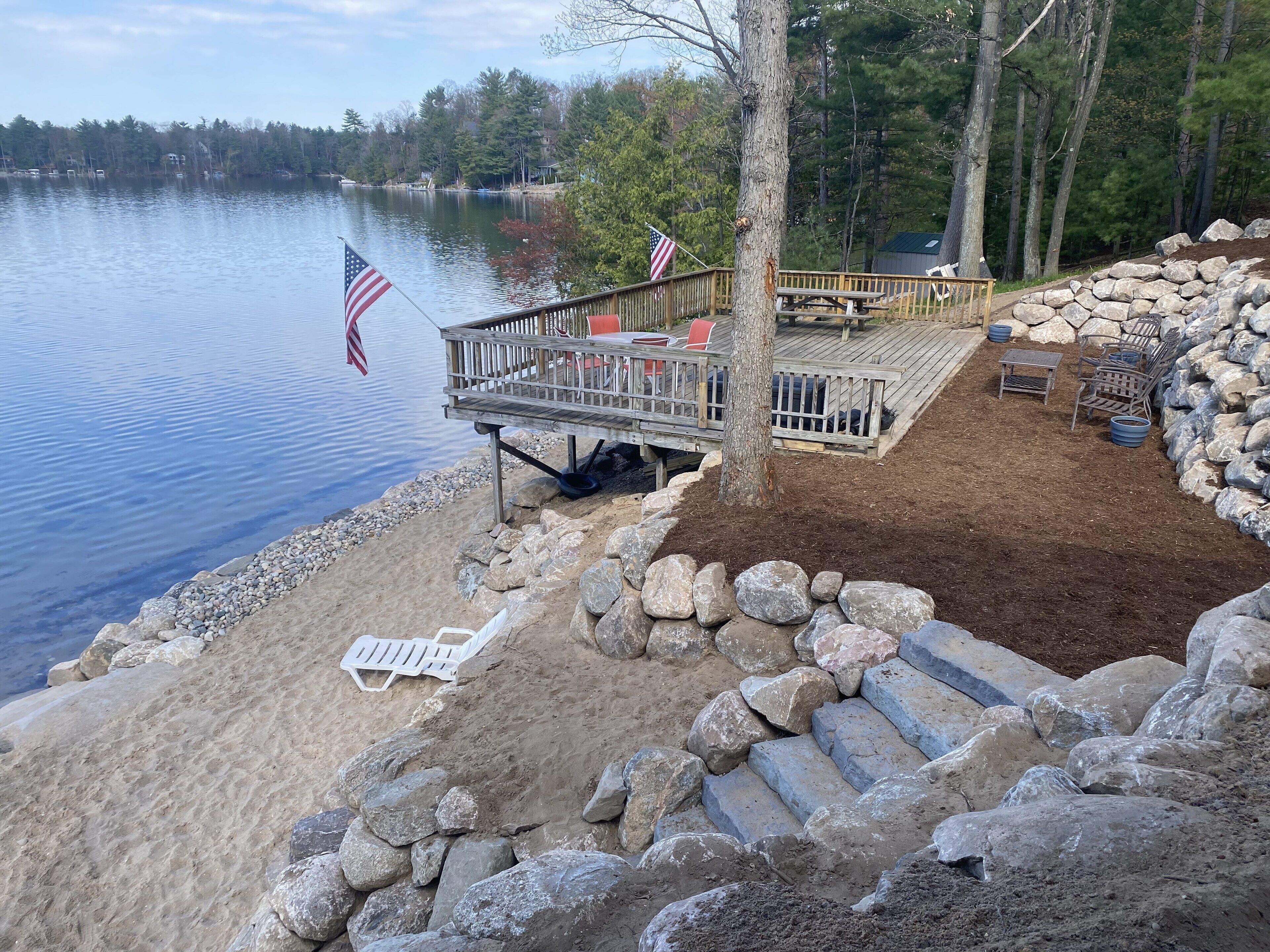 Une terrasse au bord du lac, avec une table de pique-nique et des chaises, entourée de rochers et d’une zone sablonneuse.