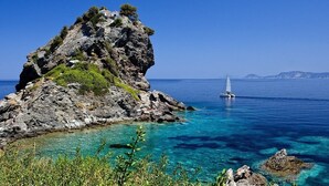 A rocky island with a building on top, clear blue waters, and a sailboat in the distance.