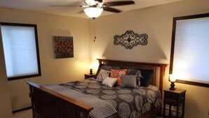 A bedroom with a wooden bed, two bedside tables with lamps, a ceiling fan, a wall-mounted artwork, and a window with blinds.