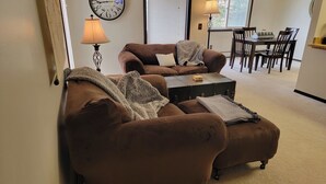 A living room with two brown sofas, a small table, a clock, and a dining area with chairs and a lamp.
