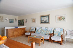 A living room with a wooden sofa, a glass coffee table, and a white chair. There is a wooden cabinet with a blue bowl on top.