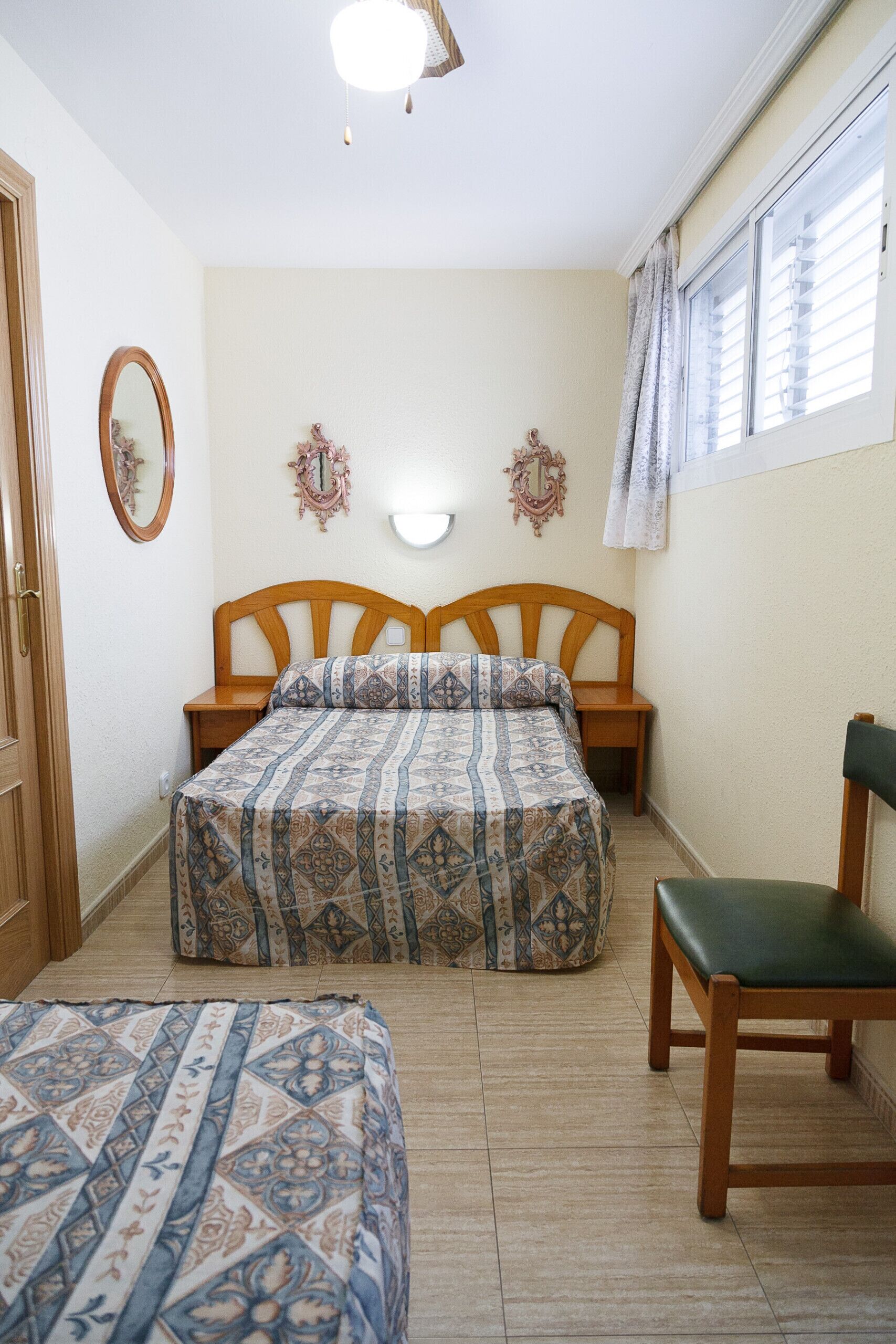 A bedroom with two beds, a wooden chair, a bedside table, and a window with curtains.
