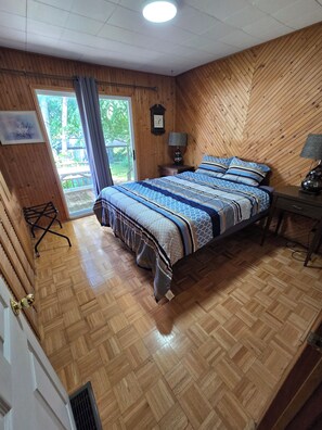 A bedroom with a wooden floor, a bed with patterned bedding, a bedside table with a lamp, a clock on the wall, and a window with curtains. - Kawartha's Home/Cottage on Buckhorn Lake (Ennismore)