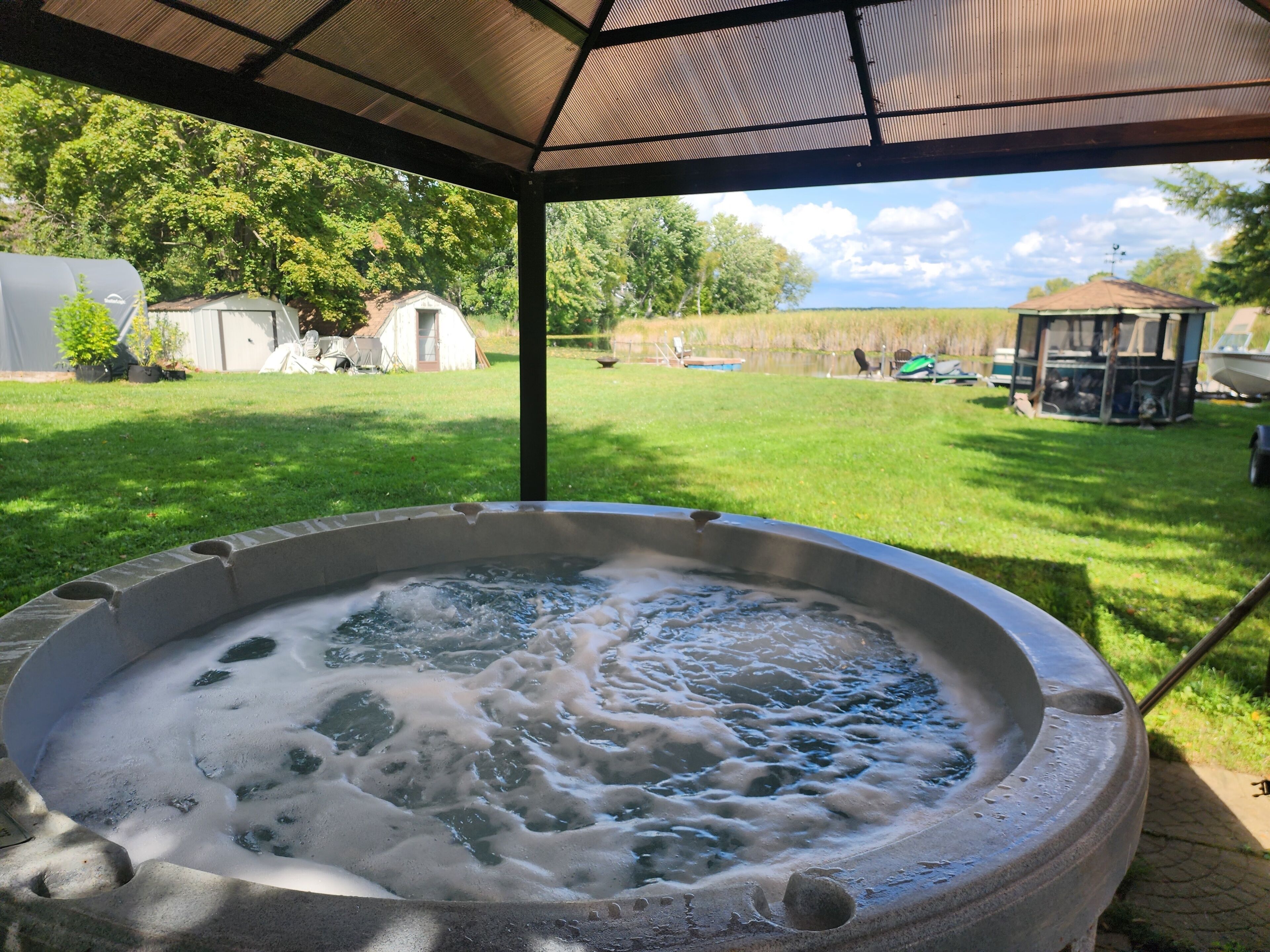 A hot tub under a covered area with a grassy yard and a gazebo in the background.