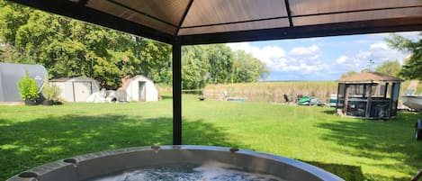 A hot tub under a covered area with a grassy yard and a gazebo in the background.