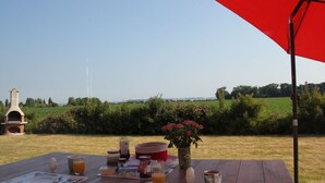 Ein Essplatz im Freien mit Frühstücksleckereien, einem roten Sonnenschirm und Blick auf ein Feld und Bäume.