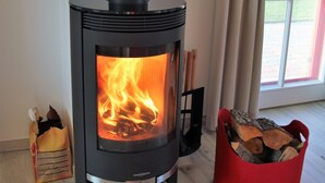 Ein Kaminofen mit sichtbarem Feuer, ein roter Holzbehälter und ein Fenster mit Vorhängen.