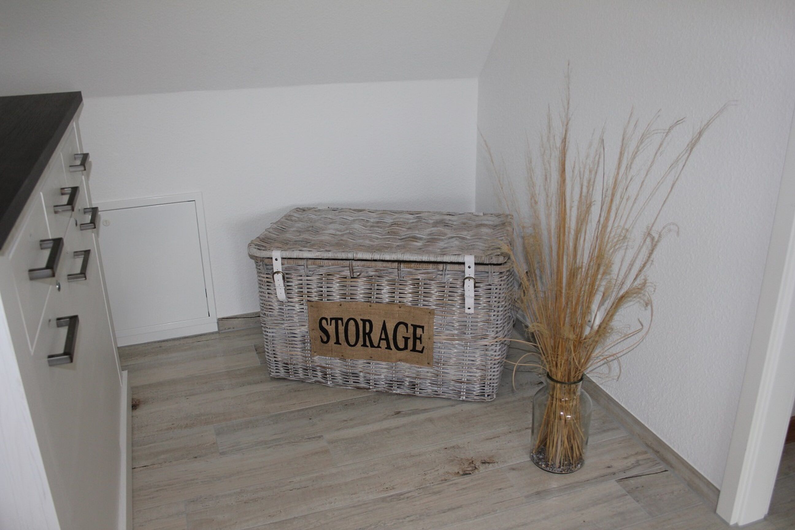 Une boîte de rangement étiquetée « RANGEMENT » est placée dans un coin, sur un plancher en bois, à côté d’un vase contenant des plantes séchées.