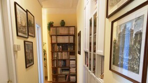 A hallway with a patterned carpet, framed pictures on the walls, a bookshelf, and a window with curtains.