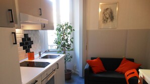 A compact kitchen with white cabinets, a sink, and a stove. There's a potted plant and a black couch with orange cushions in the living area.