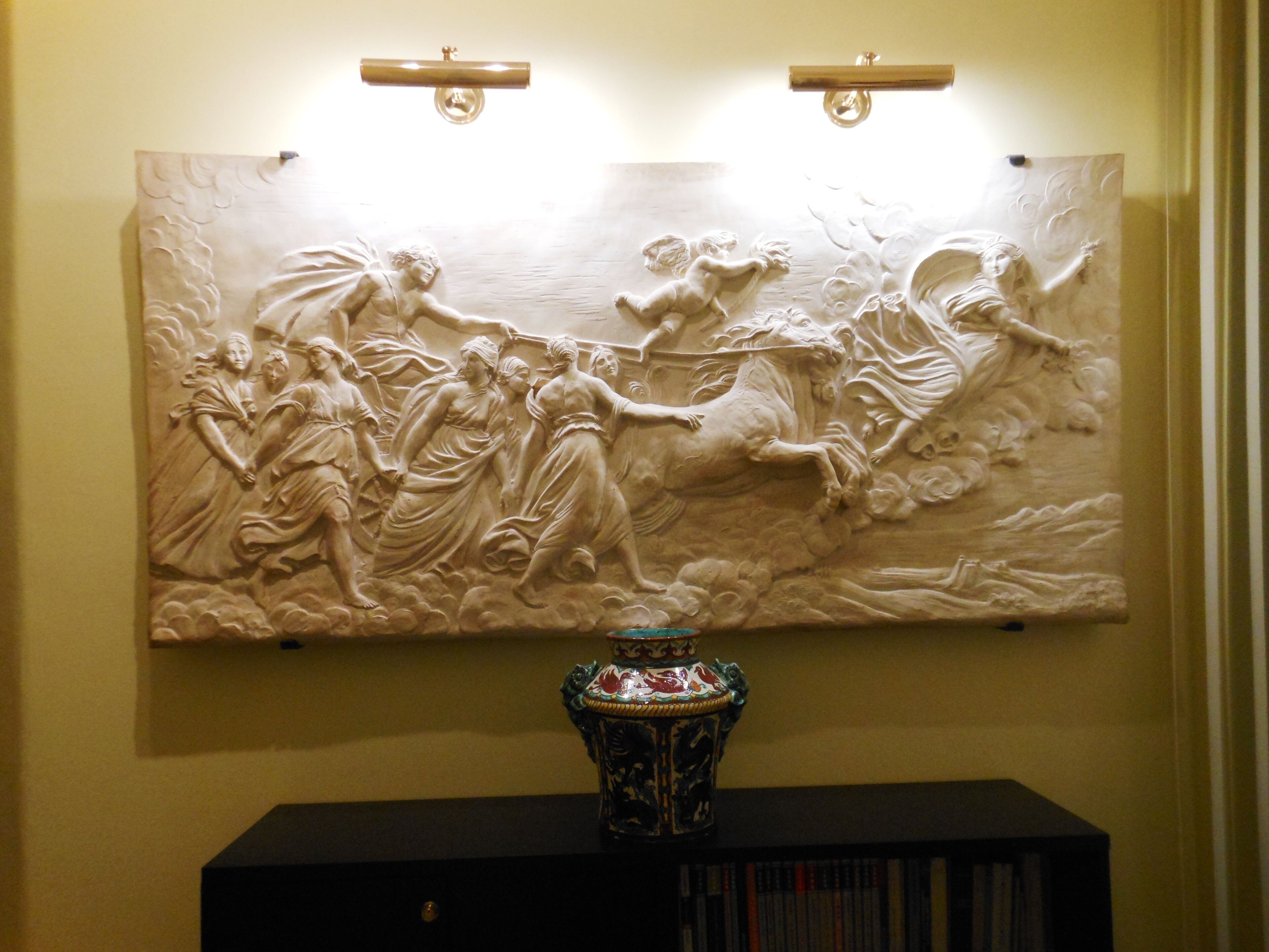 A large bas-relief sculpture on a wall, a decorative vase on a black console table, and two wall-mounted light fixtures.