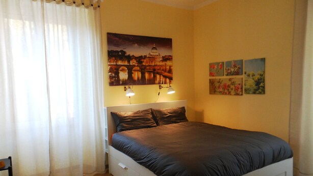 A bedroom with a bed, a nightstand, a wall-mounted lamp, and a painting of a cityscape.